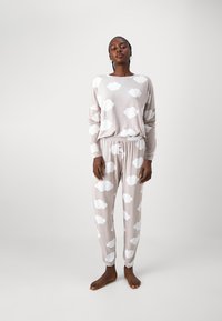 Ljusbeige loungewear-set med långärmad topp och matchande byxor, båda med vita molnmönster. Mjuk tyg, avslappnad passform.