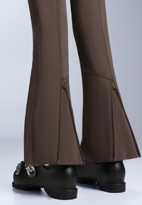 Ruskeat liehuvat housut, joissa vetoketju koristaa lahkeen reunaa, tehty sileästä, joustavasta materiaalista. Mustat puukengät, joissa metallinen solkikoriste.