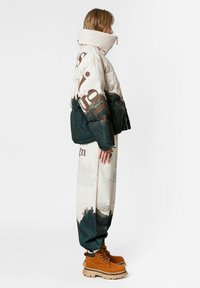 Chaqueta acolchada con cuello alto, con esquema de colores beige y verde, estampado de paisaje montañoso y grandes acentos de texto. Se lleva con pantalones estampados y botas marrones.