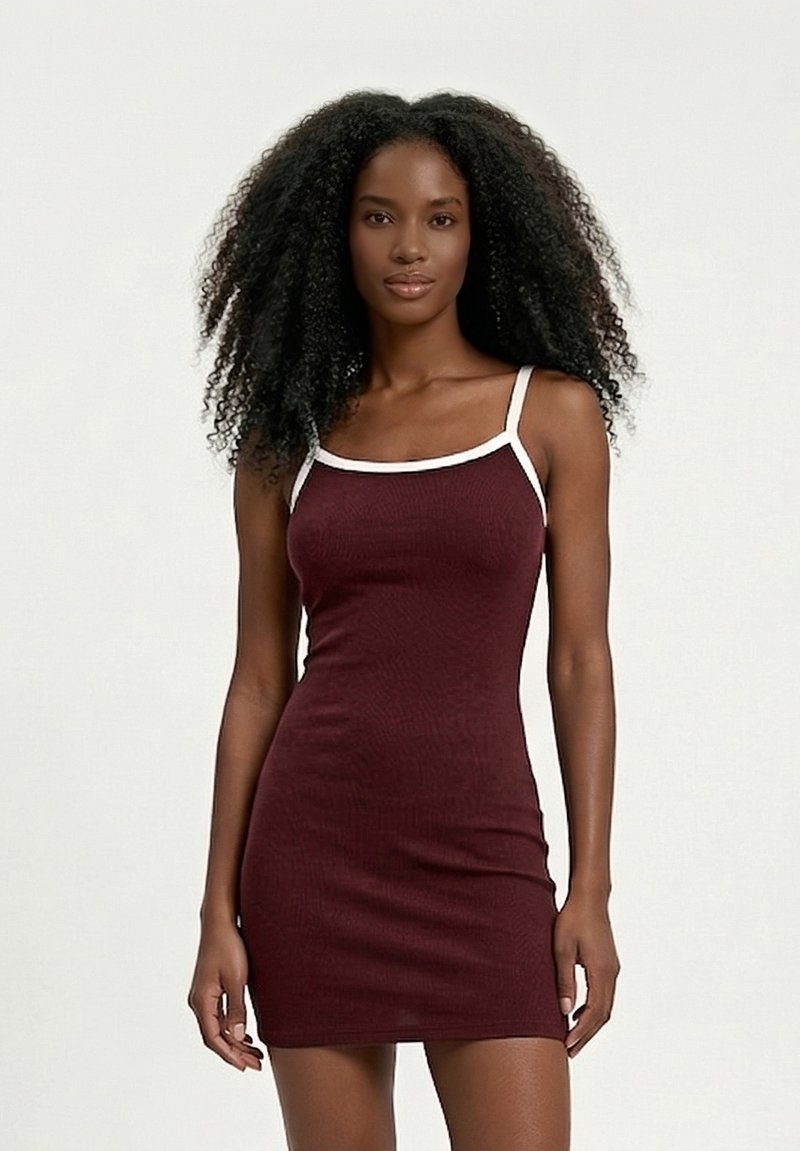 Γυναίκα με μακριά σγουρά μαλλιά που φοράει στενό αμάνικο μπορντό φόρεμα με λευκή διακόσμηση, στέκεται μπροστά σε ένα απλό φωτεινό φόντο.