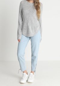 Grau gesprenkelter Strickpulli mit langen Ärmeln und abgerundetem Saum, kombiniert mit hellblauen, schmalen Jeans mit floralem Design an den Säumen. Weiße Sneakers.