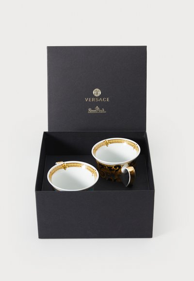 Zwei dekorative Porzellantassen mit goldfarbenen Akzenten, eine mit einem schwarzen, kunstvoll verzierten Muster, verpackt in einer schwarzen Schachtel mit goldfarbenem Logo.