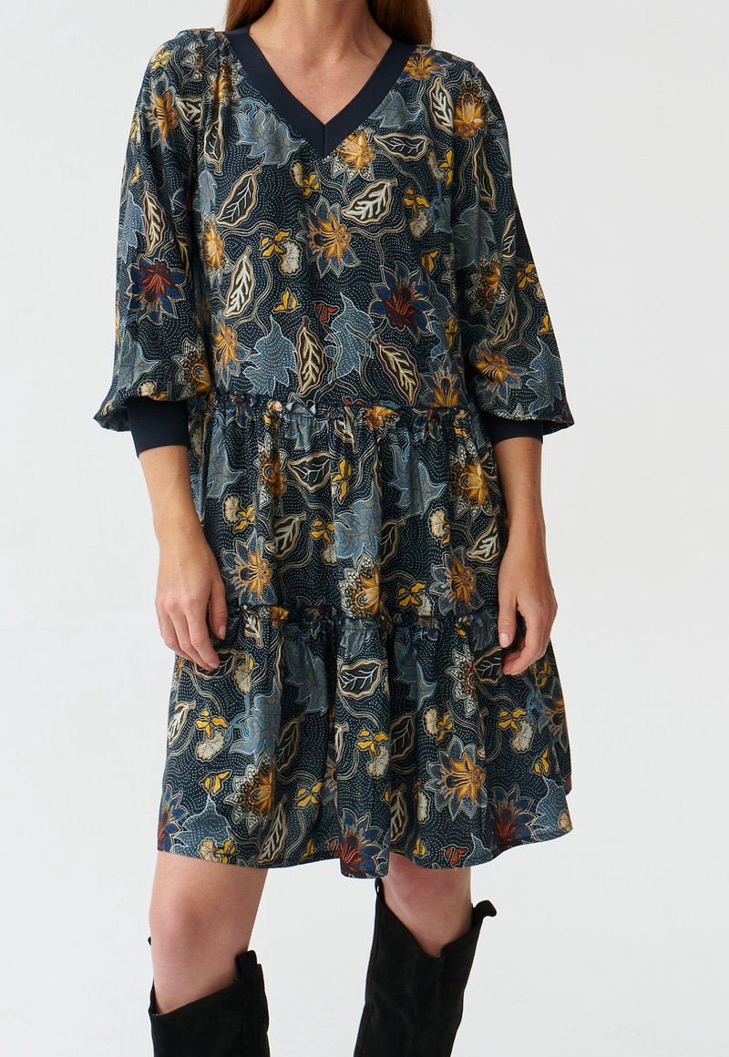 Vestido de estampado floral con escote en V azul marino, mangas de tres cuartos y falda con volantes. Hecho de tela ligera; presenta colores y patrones llamativos.