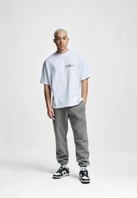 Tricou alb oversized din bumbac cu imprimeu grafic în negru și roșu; asortat cu pantaloni de trening gri și adidași alb-negru.