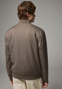 Braune Bomberjacke mit glatter Textur, geripptem Saum und Kragen sowie einem einfachen Design, von hinten vor einem neutralen Hintergrund gesehen.