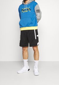 Felpa blu e grigia con accenti gialli, dotata di una tasca frontale e lettering. Abbinata a pantaloni corti in rete neri e sneaker bianche.