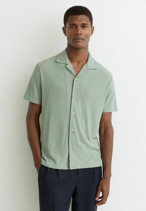 Młody mężczyzna w jasnooliwkowej koszuli z krótkim rękawem z wzorem w jodełkę i ciemnych spodniach, stojący z jedną ręką w kieszeni.