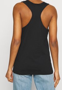 Eine Person, die ein ärmelloses schwarzes Racerback-Top und hellblaue Jeans trägt, von hinten vor einem schlichten Hintergrund gezeigt.