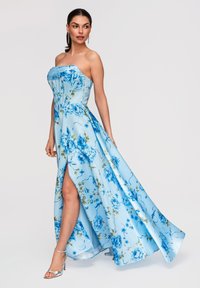 Femme portant une robe bustier bleu clair à motifs floraux avec une fente haute à la cuisse et des sandales argentées à talons hauts, posant devant un fond uni.