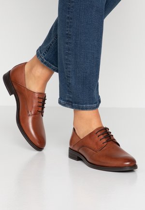 Brązowe skórzane buty z zaokrąglonym noskiem, sznurowanym zapięciem i niskim obcasem, zestawione z ciemnoniebieskimi dżinsami z podwiniętymi nogawkami. Gładka tekstura, elegancki wykończenie.