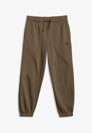 Garçon debout les mains dans les poches, portant une chemise crème à manches courtes, un pantalon vert olive et des baskets blanches sur un fond gris uni.