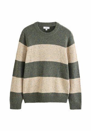 Mężczyzna ubrany w zielono-beżowy sweter w paski oraz niebieskie dżinsy stoi na trawie, patrząc w prawo w naturalnym otoczeniu na zewnątrz.
