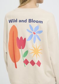 Sweat-shirt beige avec des graphiques imprimés colorés : une tulipe rouge, une fleur bleue, une feuille orange, un soleil jaune et des formes géométriques, avec le texte « Wild and Bloom ».