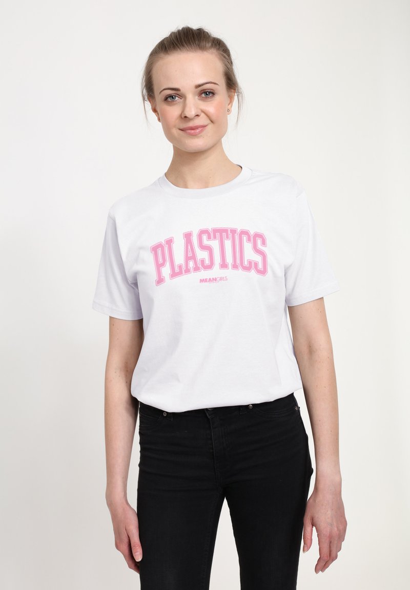 Ung kvinde iført en hvid T-shirt med pink teksten "PLASTICS MEAN GIRLS" og sorte bukser, der står mod en ensfarvet lys baggrund.