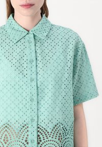Licht mintgroene shirt gemaakt van geperforeerde stof. Beschikt over een knoopsluiting aan de voorkant, een kraag en geschulpte kantdetails aan de onderkant.