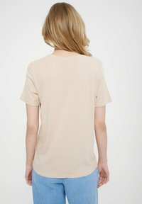 Blondyn z włosami do ramion, ubrany w gładką beżową koszulę z krótkim rękawem i jasnoniebieskie dżinsy, stojący plecami na białym tle.