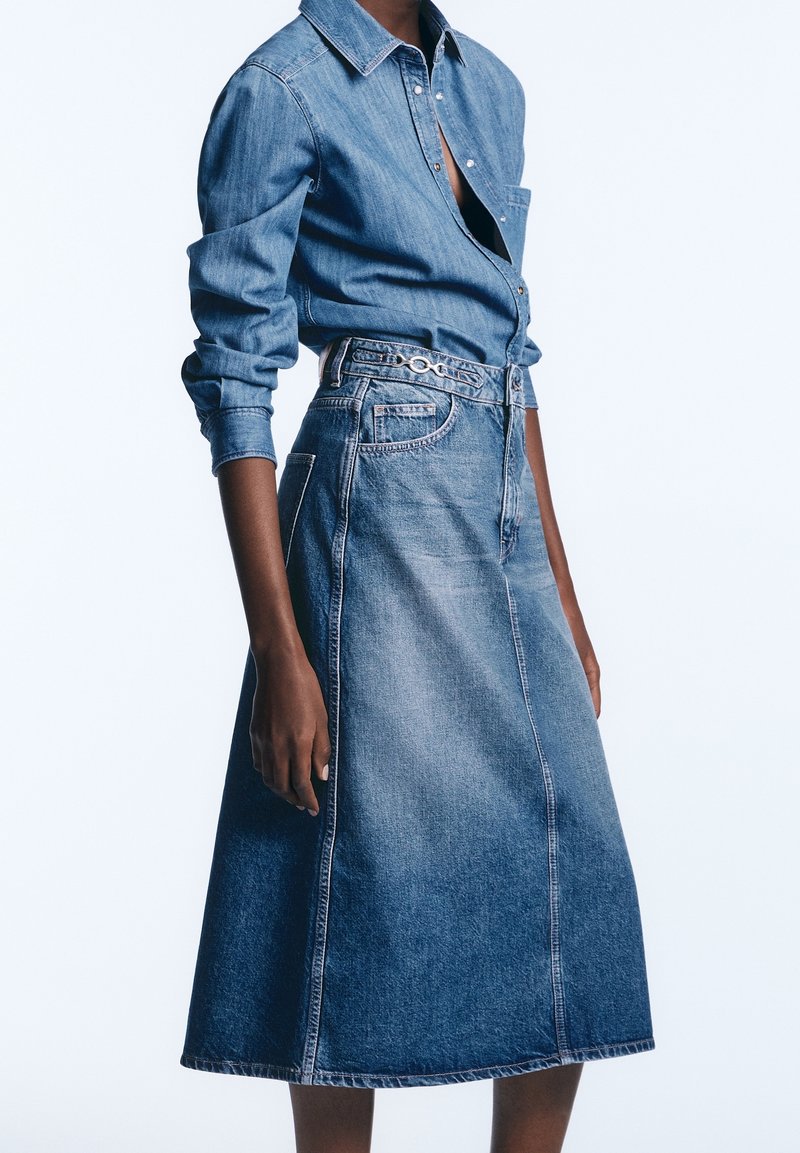 Femme portant une chemise en jean à manches longues rentrée dans une jupe en jean taille haute, coupe évasée, avec un petit détail ceinture.