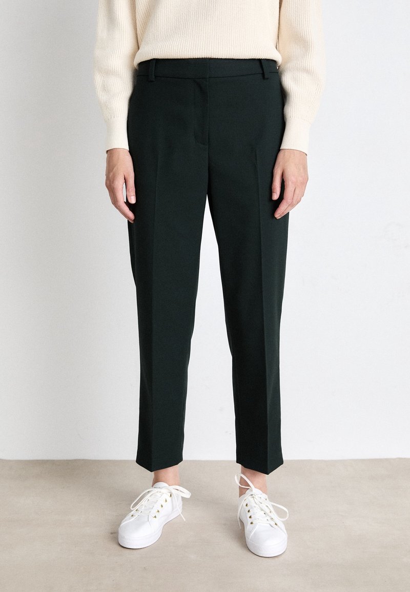 Ciemnozielone dopasowane spodnie z wąskim krojem, z wyprasowanymi zakładkami i bocznymi kieszeniami, w połączeniu z białymi sneakersami. Gładka faktura tkaniny.