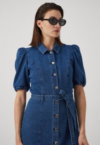 Jeansklänning med puffärmar, krage, framknappning och en knytning i midjan. Har en blå färg och silverfärgade knappar som accent.