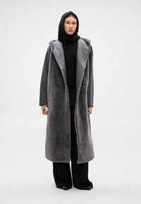 Dlhý sivý kabát s jemnou, textúrovanou povrchovou úpravou a kontrastným sivým saténovým golierom. Otvorený vpredu so zapínaním na dva gombíky, nosený cez čierne oblečenie.