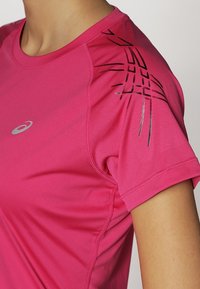 Rosa träningsskjorta med korta ärmar, prydd med svarta geometriska mönster på axlarna och en silverfärgad logotyp på bröstet. Slät tygtextur.