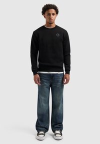 Zwarte sweatshirt met ronde hals en een klein blauw logo, gecombineerd met vervaagde blauwe wijde spijkerbroeken en zwart-witte sneakers.