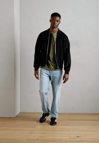 Black bomber jacket with silver zipper, olive green t-shirt, light blue jeans, and black sneakers with white accents. Wooden floor background.