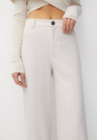 Vrouw in hoge taille, wijde crèmekleurige broek met een zwarte knop en een crèmekleurige cropped top met lange mouwen van ribstof, tegen een effen achtergrond.
