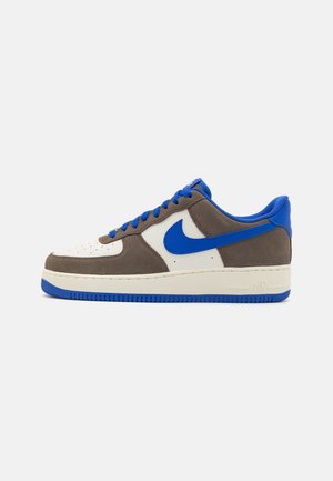 Baskets basses avec panneaux en daim beige et marron, lacets bleus, logo Nike bleu et semelle en caoutchouc bleue, sur fond blanc.