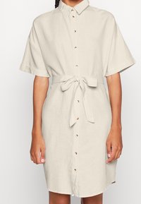 Vestito in lino beige chiaro con maniche corte, chiusura frontale a bottoni e cintura annodata in vita. Presenta bottoni in tonalità dorata e una vestibilità comoda.