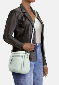 Borsa a tracolla in pelle verde menta chiaro con spallaccio regolabile, tasca frontale con zip e texture liscia, indossata con una giacca di pelle marrone e jeans blu.