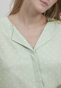 Šviesiai žalia blūzė su baltais taškeliais, turinti išpjautą apykaklę ir mygtukų užsegimą priekyje, pagaminta iš lygaus, minkšto audinio.