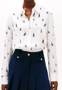Chemise blanche à manches longues avec un motif de voilier, dotée d'un col pointu et d'une patte de boutonnage, associée à une jupe plissée navy avec des boutons dorés.