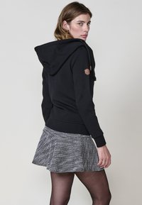 Sudadera negra con un patrón texturizado, capucha ajustable con cordón y un pequeño parche de cuero en la manga izquierda; combinada con una falda de tejido gris.