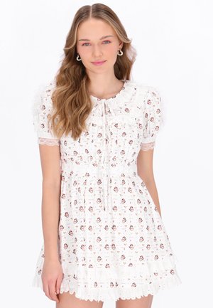 Młoda kobieta o długich falowanych włosach ubrana w białą kwiecistą krótką sukienkę z bufkami i koronkowym wykończeniem, stojąca na jednolitym białym tle.