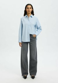 Camicia azzurro chiaro con maniche lunghe, abbinata a jeans grigi scuri a gamba larga e décolleté neri a punta. Design semplice, colori solidi.