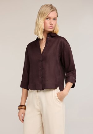 Blonde Frau trägt ein dunkelbraunes Hemd mit Knöpfen und hochgekrempelten Ärmeln sowie hellbeige Hose, eine Hand in der Tasche, neutraler Hintergrund.