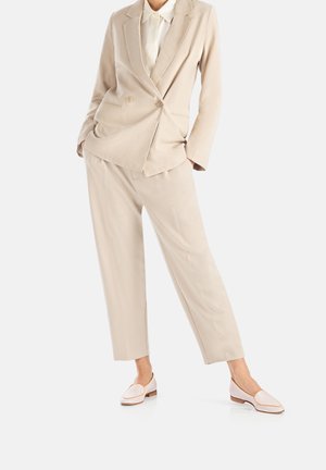 Beige double-breasted suit jacket and matching trousers, featuring a relaxed fit. White shirt underneath, paired with light pink loafers.