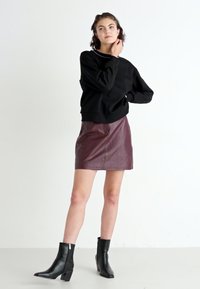 Felpa nera con orlo a costine, abbinata a una mini gonna in pelle bordeaux. Contrasto di texture trasparente con stivaletti neri alla caviglia, caratterizzati da un tacco geometrico.