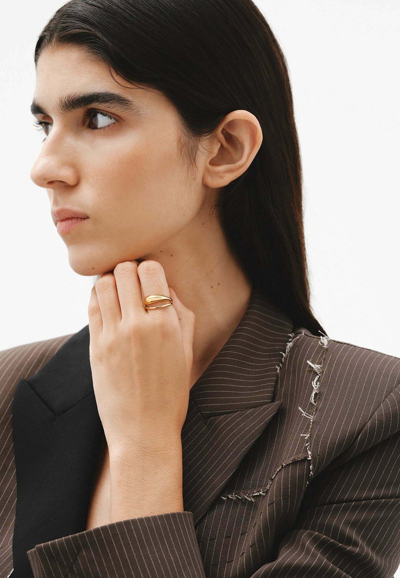 Anillo de oro con un diseño suave y entrelazado, llevado en una mano que descansa cerca del mentón frente a un fondo de un blazer marrón a rayas.