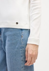 Witte langmouwtop met een ribbelstructuur, voorzien van een kleine ronde zwarte hardwareaccent aan de zoom, gecombineerd met lichtblauwe spijkerbroeken.