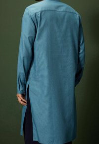 Man in een lange, teal gekleurde shirt met een textuur en lange mouwen, knoopmanchetten en zijsplits. Hij staat tegen een effen donkergroene achtergrond.