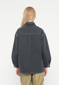 Chemise surdimensionnée gris foncé avec un col, des poignets boutonnés et un détail de pli à l'arrière. Texture lisse et coupe décontractée.