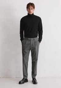 Schwarzer Rollkragenpullover kombiniert mit grauen strukturierten Hosen; das Modell steht mit den Händen in den Taschen; schwarze Schuhe runden das Outfit ab.