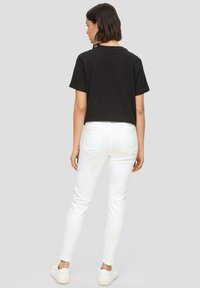 Schwarzes kurzes T-Shirt und weiße schmal geschnittene Jeans. Das T-Shirt hat einen runden Ausschnitt; die Jeans verfügen über Gesäßtaschen. Weiße Sneaker vervollständigen das Outfit.