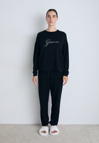 Frau mit dunklem Haar in schwarzem Loungewear-Sweatshirt und -Hosen, trägt weiße, kuschelige Pantoffeln mit offenen Zehen, steht an einer einfachen Wand auf einem Teppich.