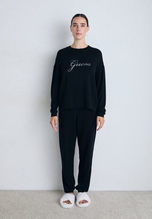 Kvinna med mörkt hår i svart loungewear, sweatshirt och byxor, bärande vita fluffiga tofflor med öppen tå, stående mot en enkel vägg på heltäckningsmatta.