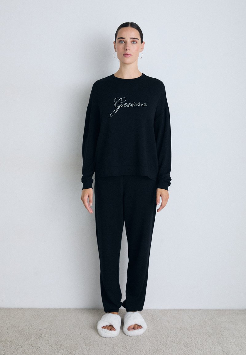Frau mit dunklem Haar in schwarzem Loungewear-Sweatshirt und -Hosen, trägt weiße, kuschelige Pantoffeln mit offenen Zehen, steht an einer einfachen Wand auf einem Teppich.