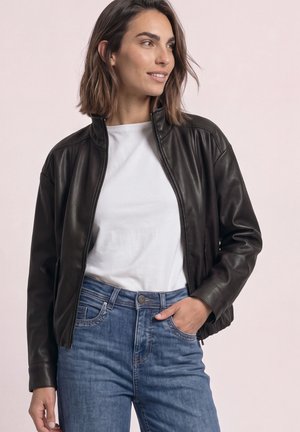 Mujer con una chaqueta de cuero negra sobre una camisa blanca y vaqueros azules, de pie con una mano en el bolsillo, mirando hacia un lado y sonriendo.