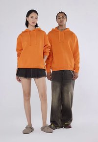 Två modeller bär överdimensionerade orangea hoodies med framfickor och dragsnören. Den kvinnliga modellen kombinerar sin hoodie med en svart kjol; den manliga bär bylsiga byxor.